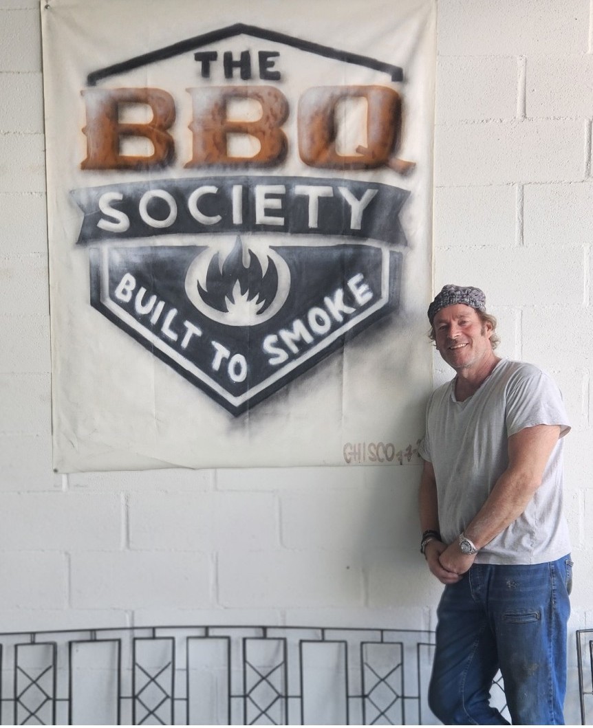 Chrétien Schatorie en el taller de The BBQ Society en La Cistérniga, junto al cartel pintado a mano de la marca
